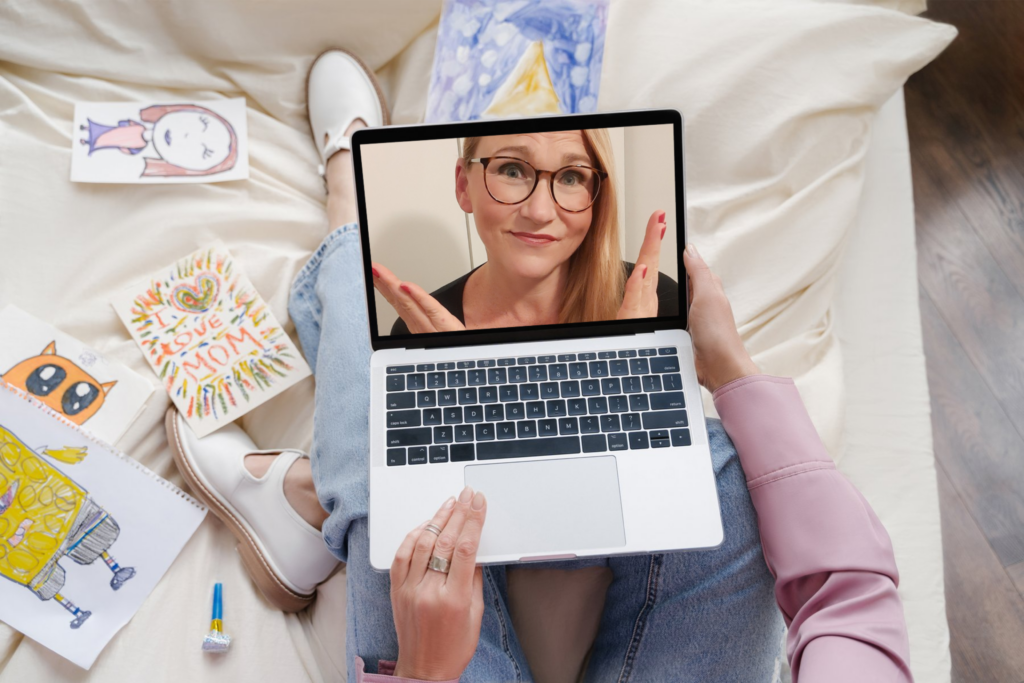 Mother sitting on her bed with her laptop in front of her. On the bed are drawings from her children. The screen is showing Barbara, the founder of #VirtualSpaceHero.